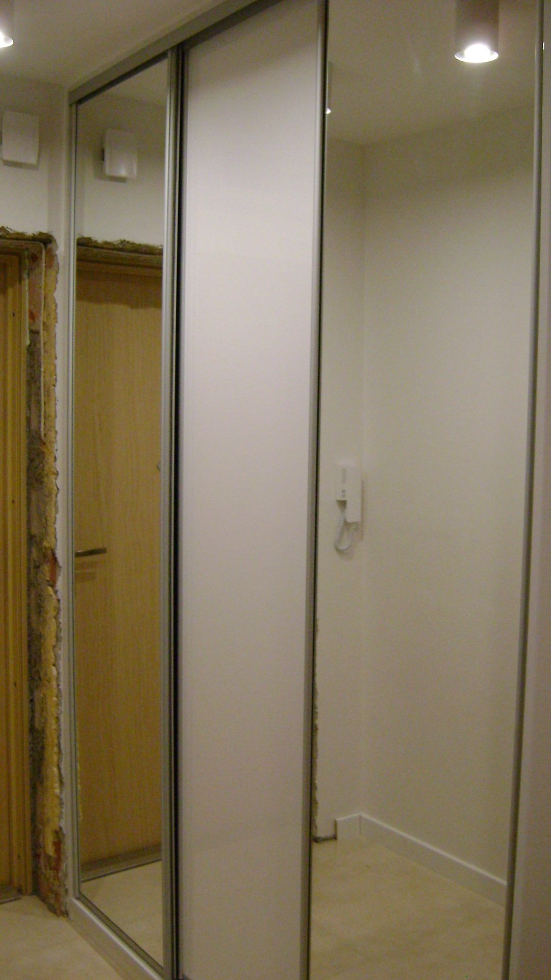 Szafa przesuwna z białymi frontami i lustrzanym panelem w przedpokoju, widoczny fragment ościeżnicy drzwi i domofon na ścianie.