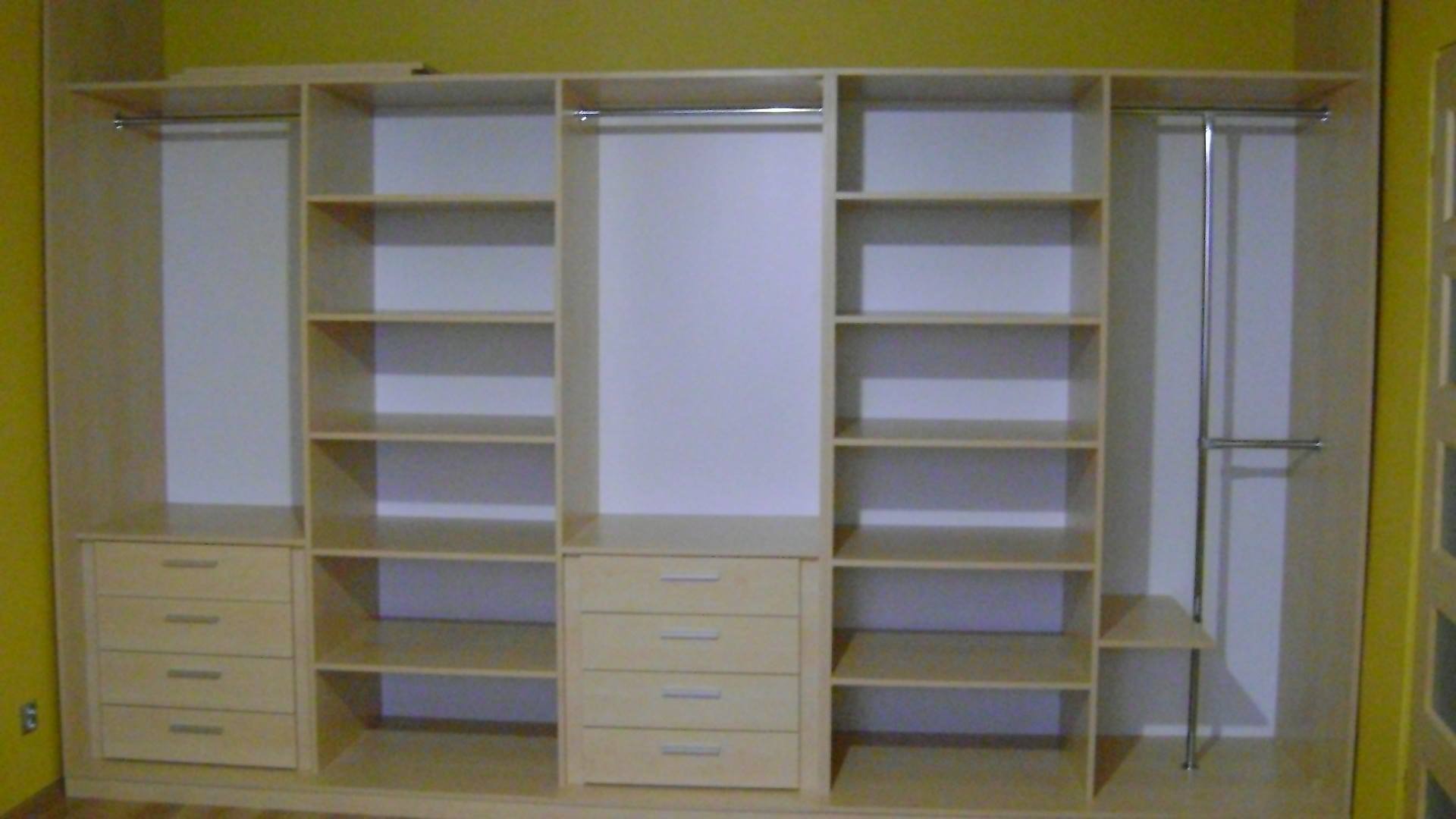 Jasna, drewniana konstrukcja szafy z szufladami i półkami, z drążkami na ubrania, na tle żółtej ściany.