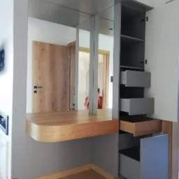 Szara szafa wnękowa z lustrami i drewnianym blatem, otwarte szuflady z drewnianym wnętrzem, widoczne drzwi w tle.