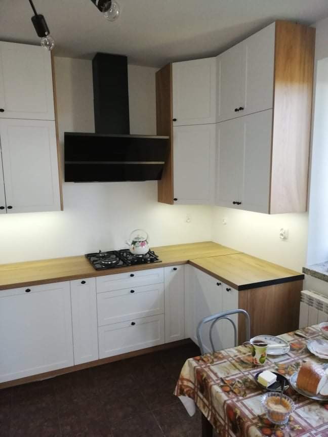 Jasna kuchnia z białymi szafkami i drewnianym blatem, czarny okap nad kuchenką gazową, stół z nakryciem i śniadaniem w tle.