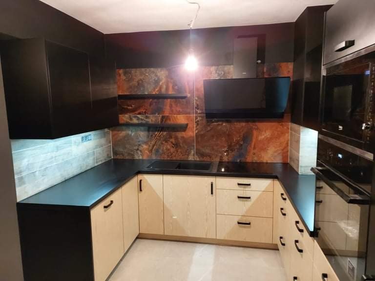 Nowoczesna kuchnia z jasnymi frontami szafek i czarnym blatem, wykończona okładziną ścienną imitującą marmur w odcieniach brązu i szarości, oświetlona punktowo, z widocznym piekarnikiem w czarnej...