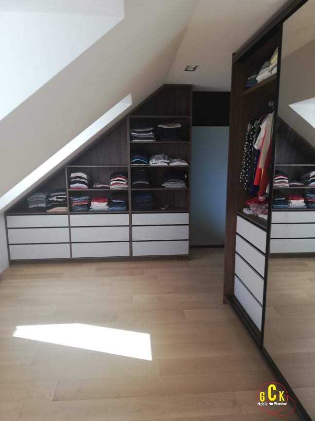 Szafa wnękowa z lustrzanymi drzwiami przesuwnymi, dopasowana do skosu dachu, z białymi szufladami i ciemnymi półkami na ubrania, na jasnej drewnianej podłodze z odbiciem światła.