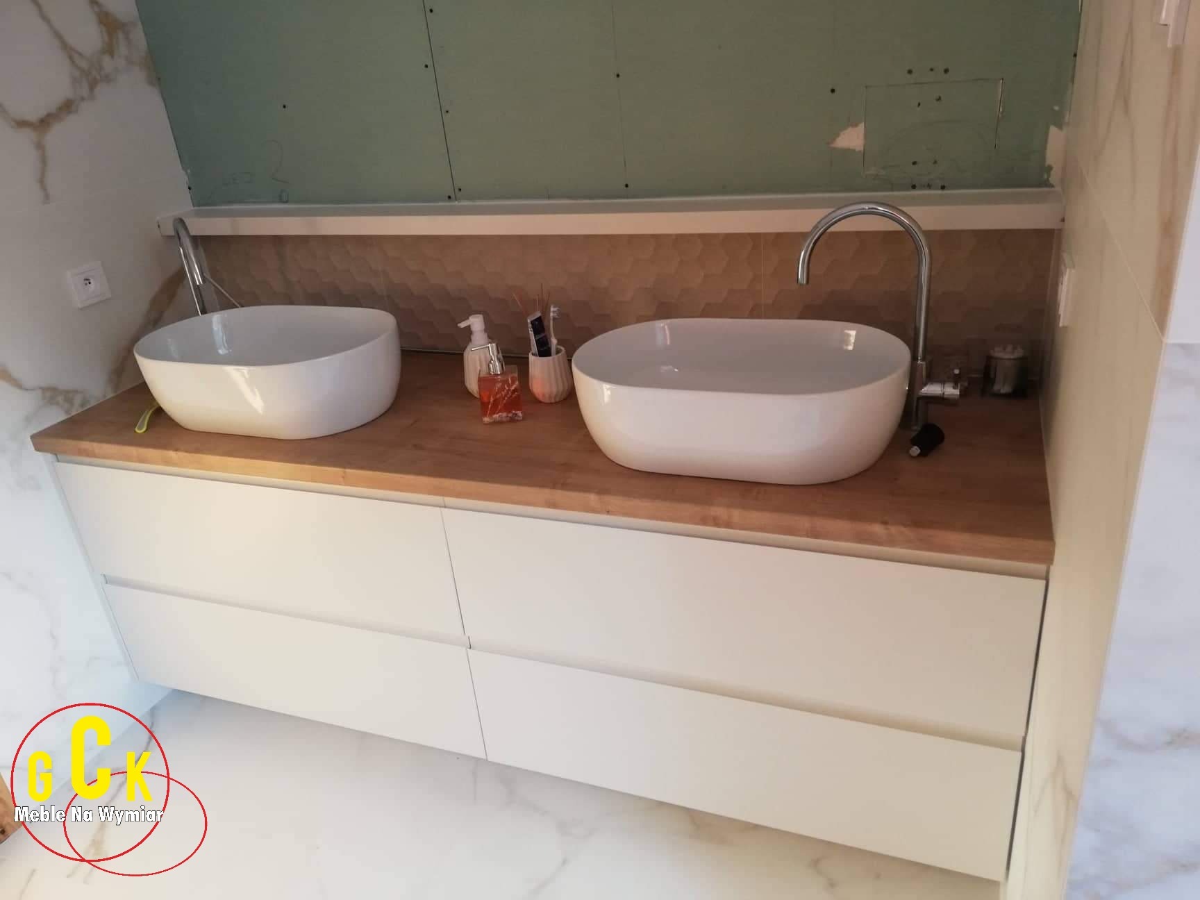Łazienka z dwiema owalnymi umywalkami nablatowymi na drewnianym blacie i białymi szafkami z szufladami. W tle widoczna ściana z dekoracyjnymi panelami i fragment ściany w trakcie wykończenia.