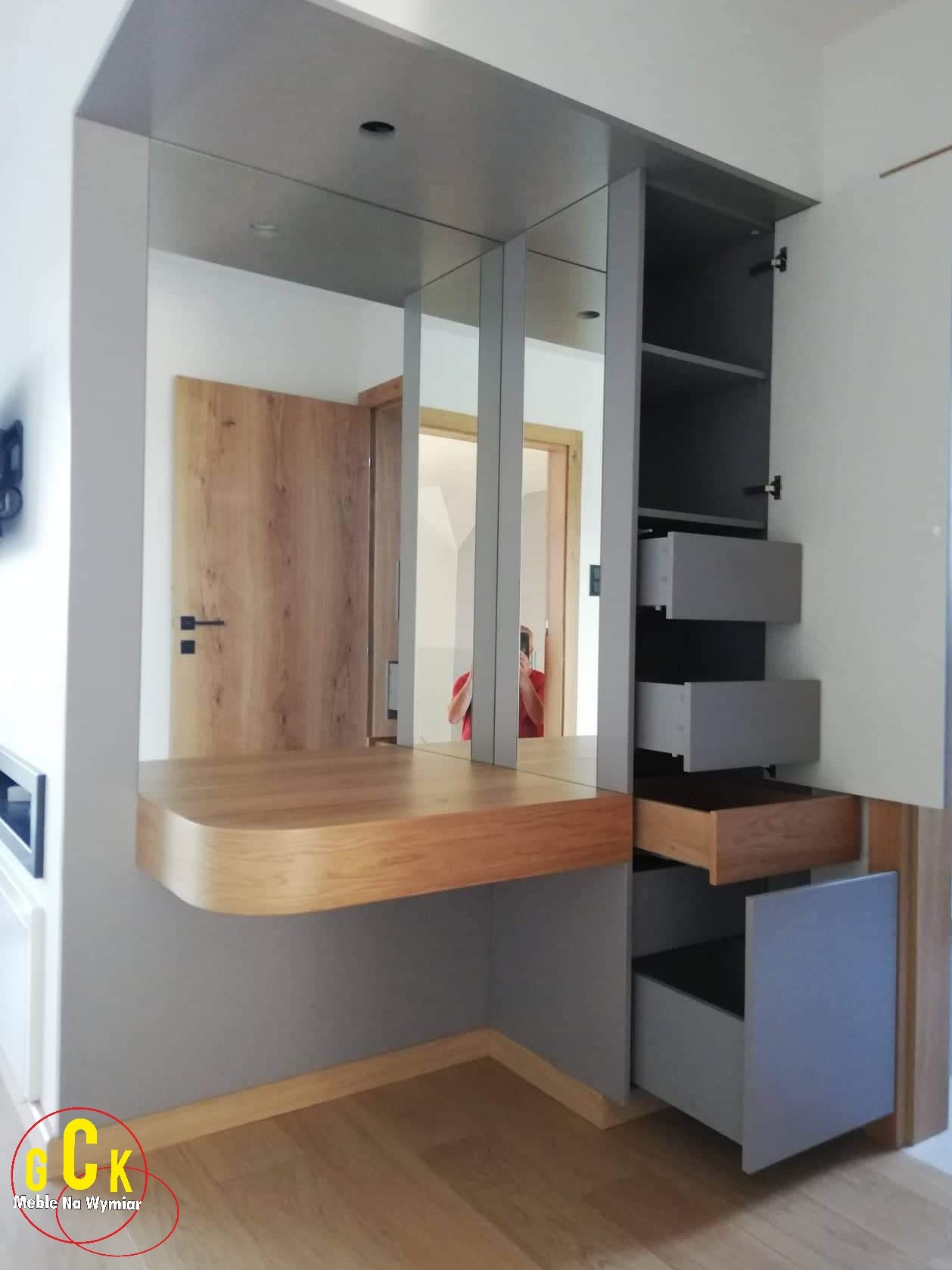 Szara szafa wnękowa z lustrami i drewnianym blatem, otwarte szuflady z drewnianym wnętrzem, widoczne drzwi w tle.