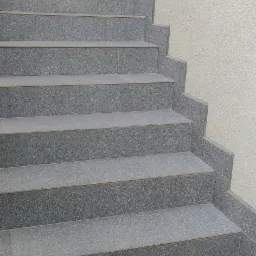 Schody wyłożone szarą, kwadratową terakotą z fugami, prowadzące do góry, z fragmentem jasnej ściany w tle.