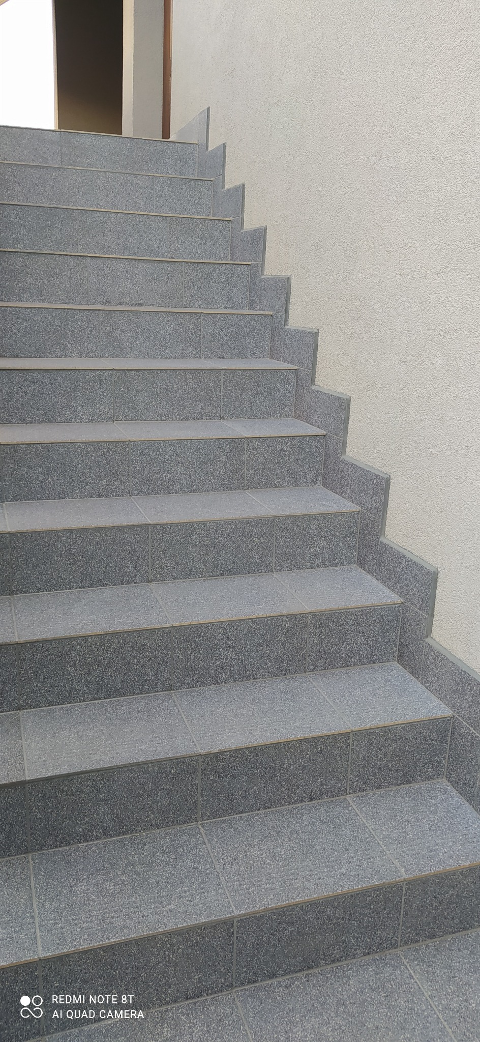 Schody wyłożone szarą, kwadratową terakotą z fugami, prowadzące do góry, z fragmentem jasnej ściany w tle.