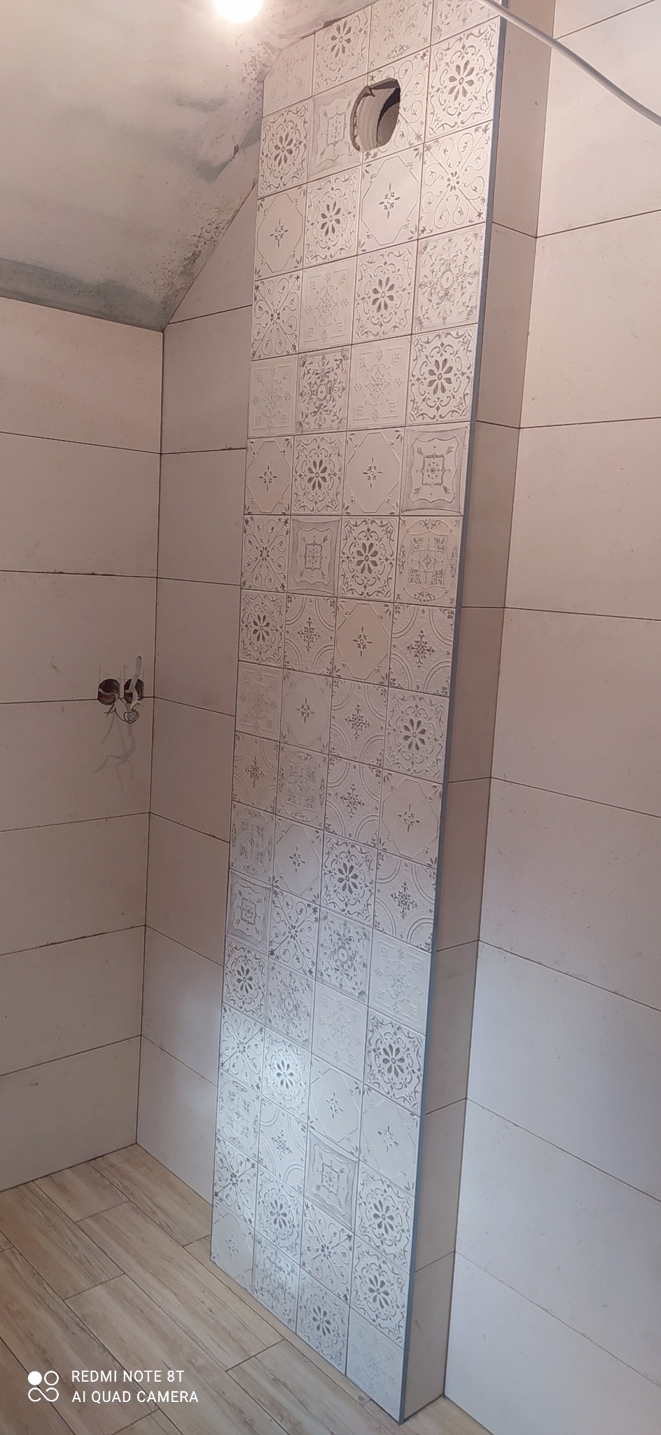 Fragment łazienki w trakcie remontu; widoczna ściana z ułożonymi płytkami ceramicznymi w biało-szare wzory, z otworem na instalację, oraz fragment podłogi z paneli i ściana z widocznymi przewodami.