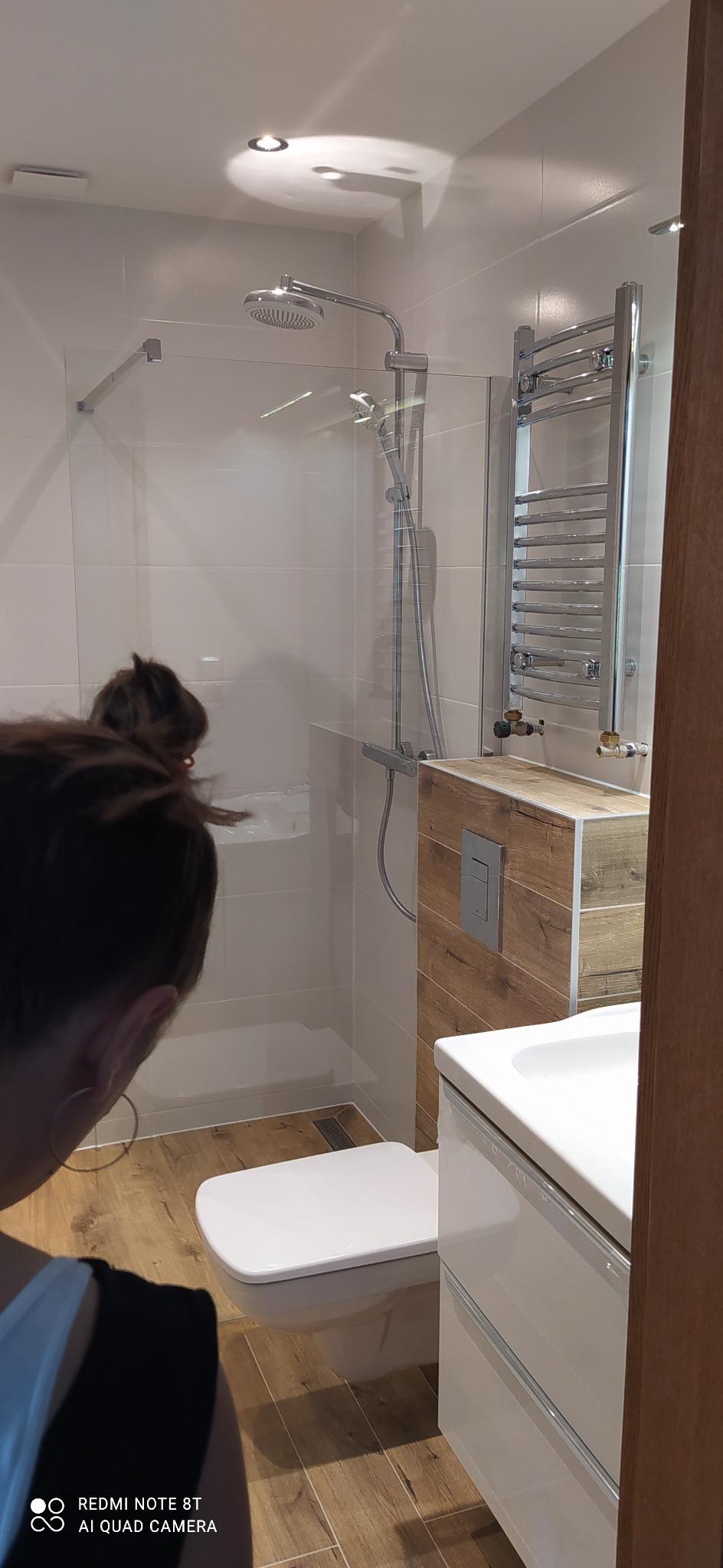 Nowoczesna łazienka z kabiną prysznicową typu walk-in, grzejnikiem drabinkowym i umywalką z szafką w odcieniach bieli i drewna, widoczna osoba w odbiciu.