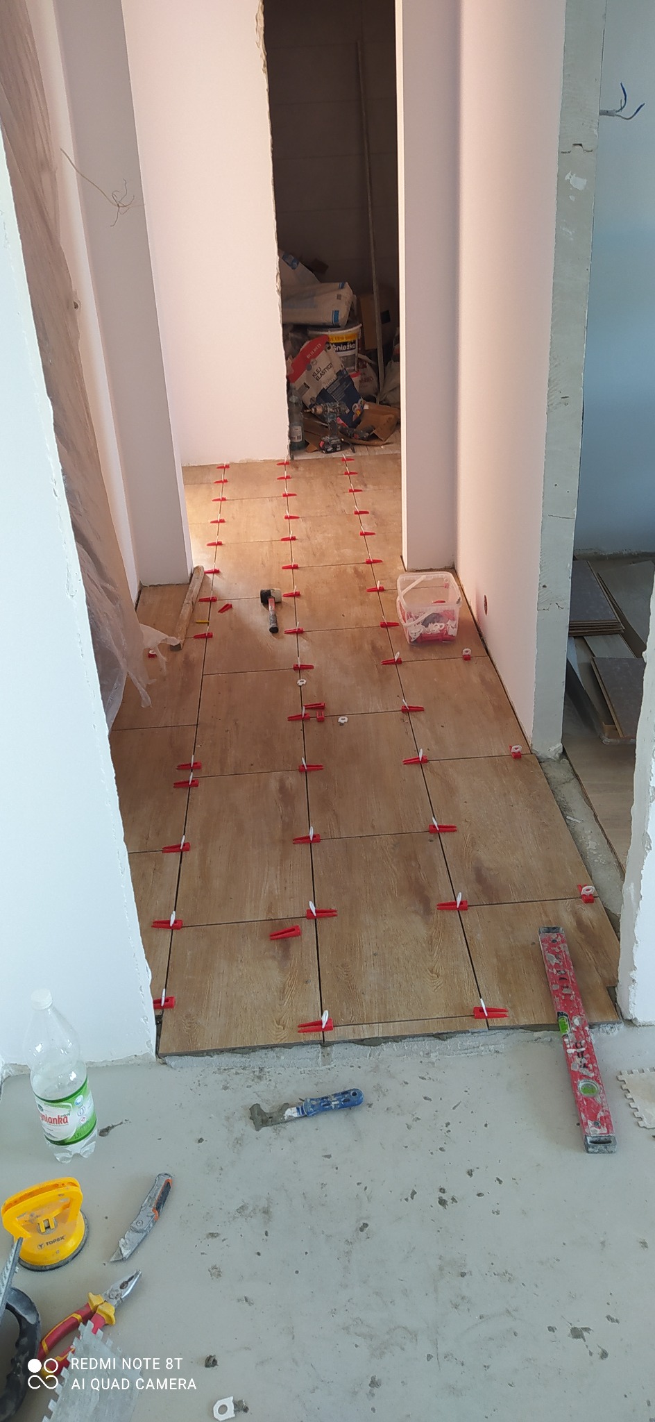 Układanie płytek podłogowych imitujących drewno w korytarzu, z użyciem systemu poziomowania płytek z czerwonymi klipsami i klinami, widoczne narzędzia pracy i poziomica.