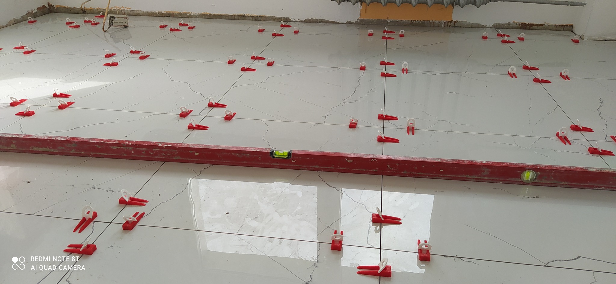 Położone białe płytki podłogowe z czerwonymi klinami poziomującymi i poziomicą w trakcie układania.