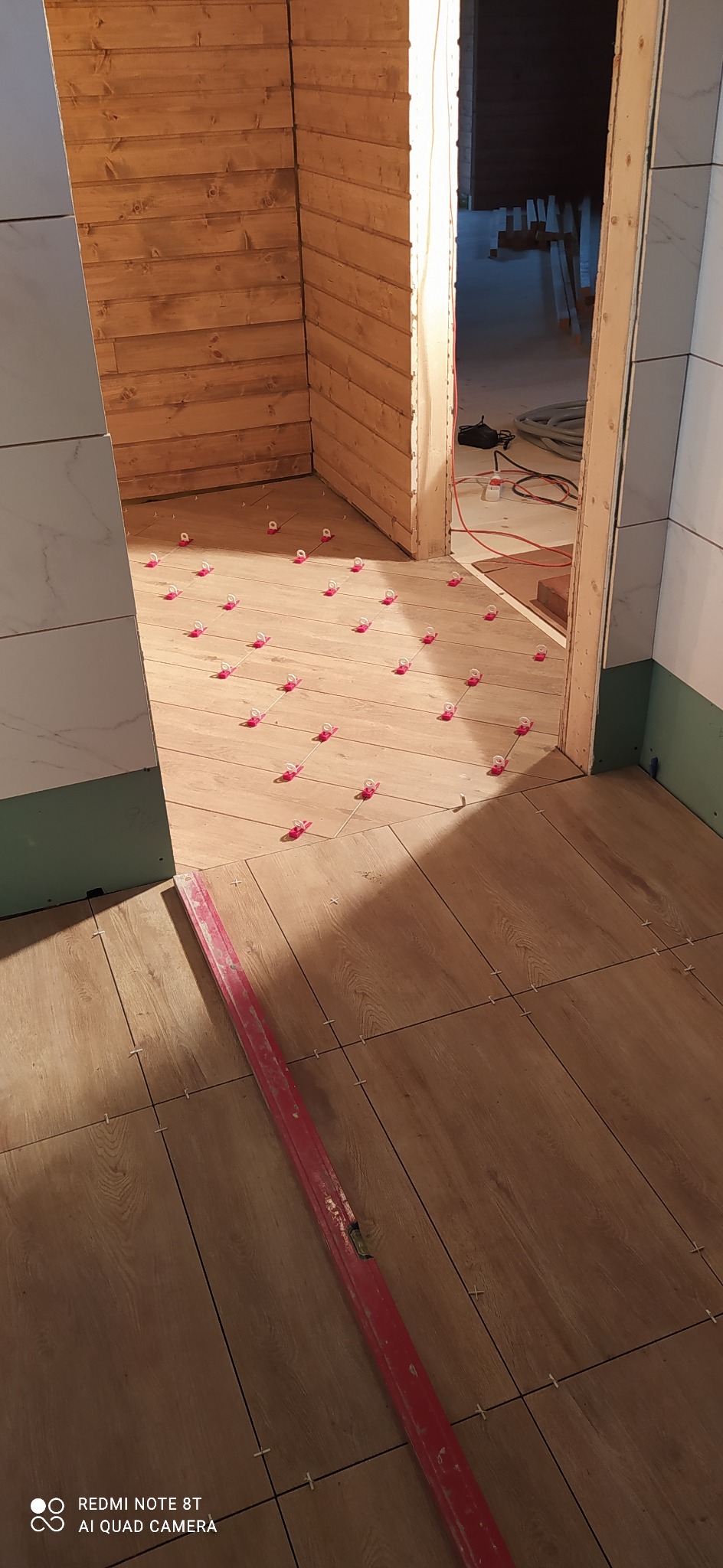 Układanie płytek imitujących drewno w łazience, widoczne krzyżyki dystansowe i poziomica czerwona, ściany wykończone panelami drewnianymi i płytami gipsowo-kartonowymi.