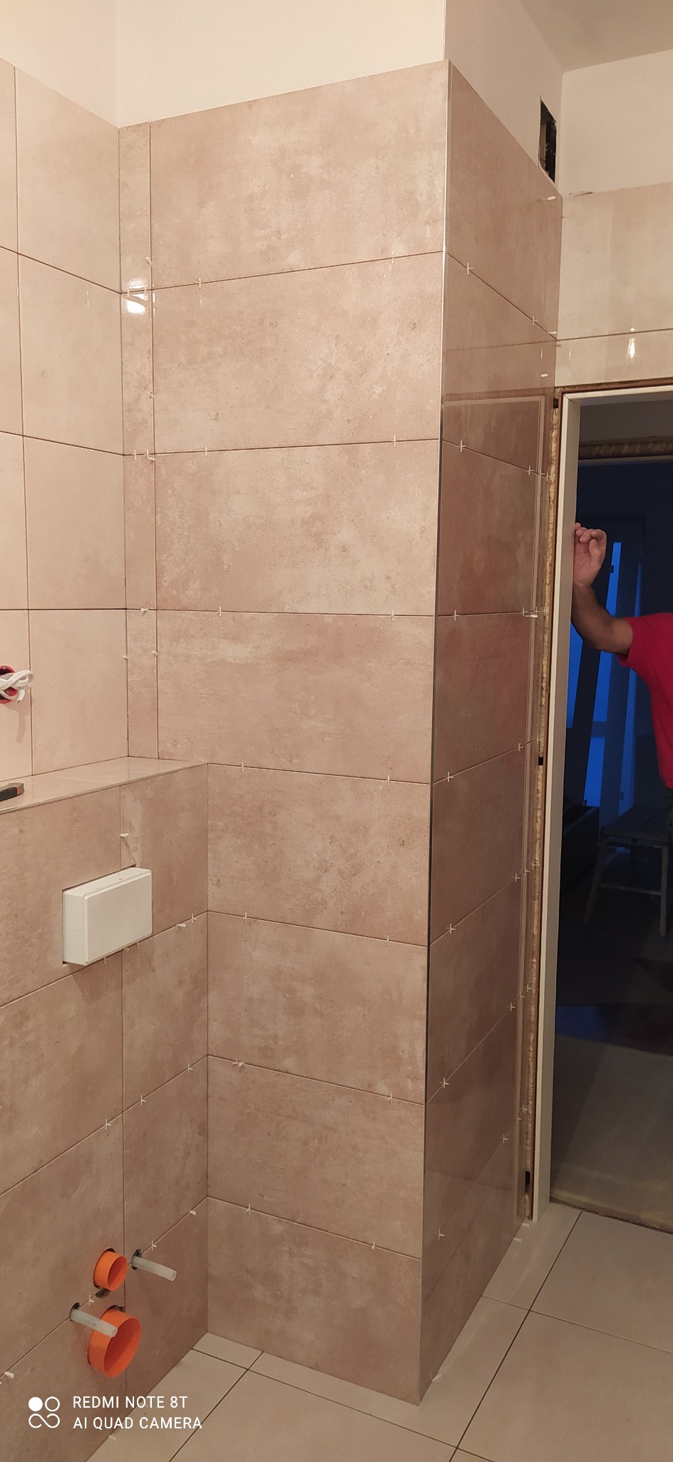 Świeżo ułożone beżowe płytki w łazience, z widocznymi krzyżykami dystansowymi, biała spłuczka podtynkowa i pomarańczowe rury odpływowe.