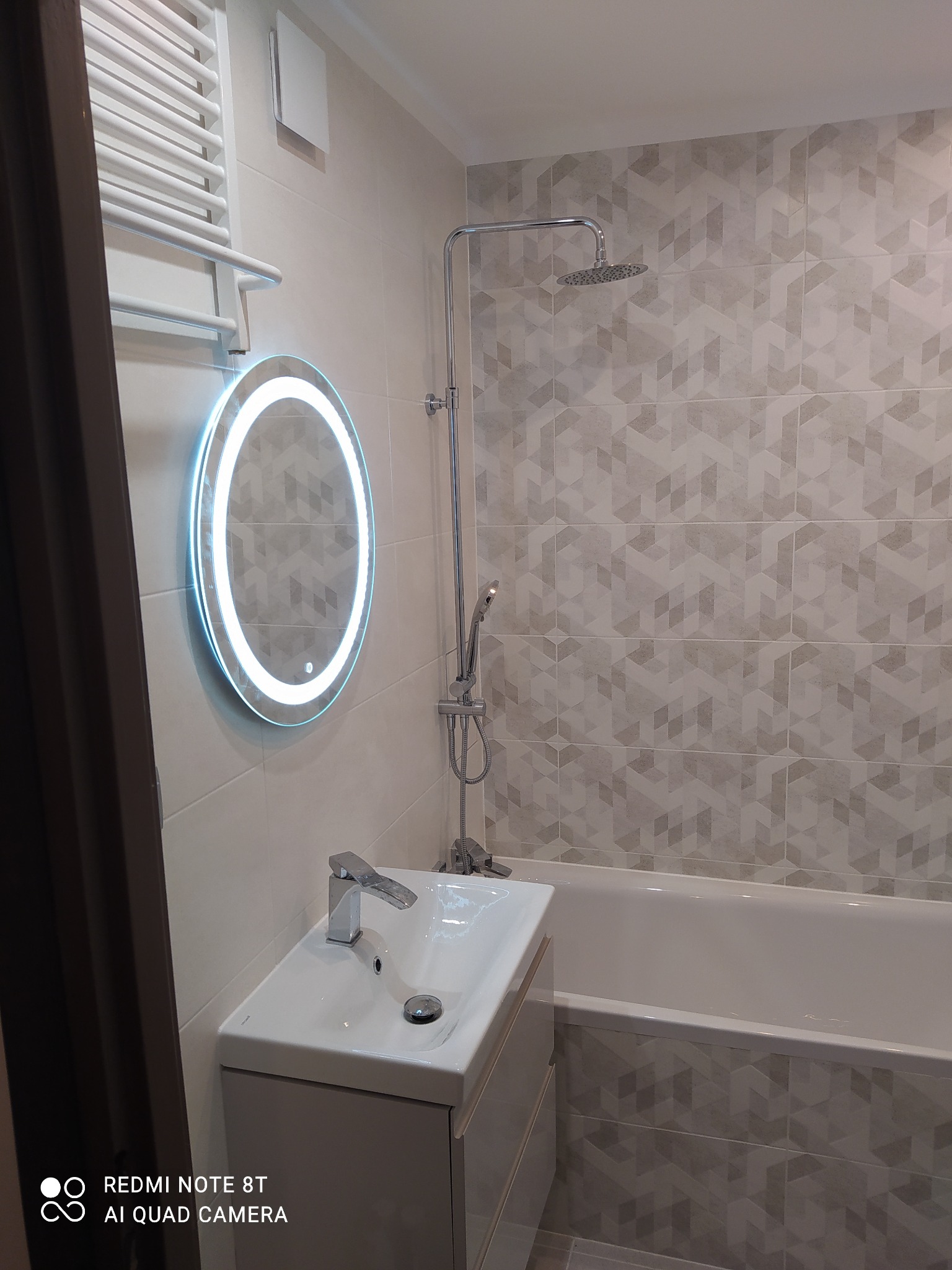 Nowoczesna łazienka z umywalką nablatową, podświetlanym lustrem LED, baterią chromowaną, prysznicem deszczownicą i geometrycznymi płytkami w odcieniach szarości.