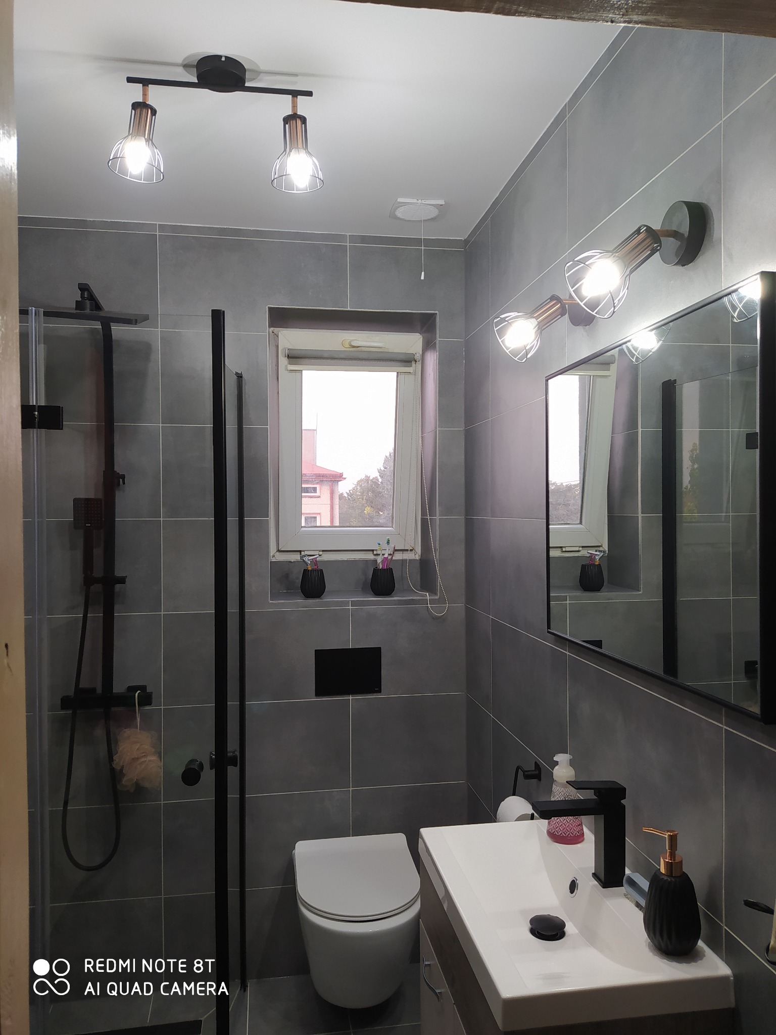 Nowoczesna łazienka w odcieniach szarości z czarnymi akcentami, widoczna kabina prysznicowa z deszczownicą, umywalka z czarną armaturą i dozownikiem mydła, wisząca toaleta, oświetlenie punktowe...