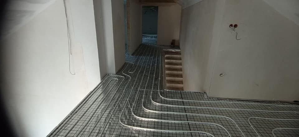 Rozłożona instalacja ogrzewania podłogowego na siatce w surowym wnętrzu, widoczne białe rury grzewcze i przygotowane schody w głębi pomieszczenia przed wylewką.