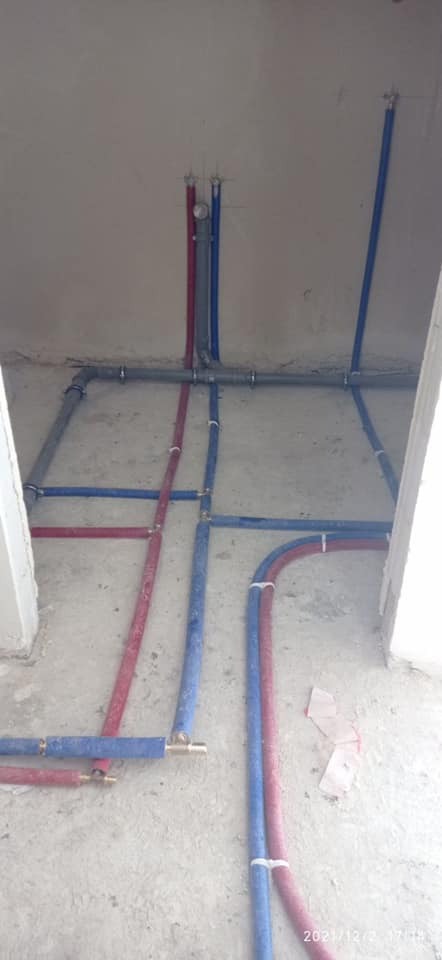 Instalacja wodno-kanalizacyjna w surowym stanie, widoczne rury niebieskie i czerwone ułożone na podłodze i przymocowane do ściany, złącza mosiężne, brak wykończenia ścian i podłóg.