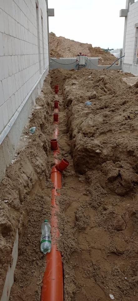 Pomarańczowe rury kanalizacyjne ułożone w wykopie ziemnym wzdłuż fundamentów budynku w trakcie budowy. Widoczne złączki i kształtki. Na dnie wykopu leży pusta butelka PET.