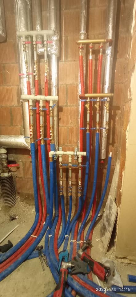 Rozdzielacz instalacji wodnej z rurami PEX w kolorze niebieskim i czerwonym, zamontowany na ścianie z cegły, widoczne narzędzia i rękawice robocze na podłodze.