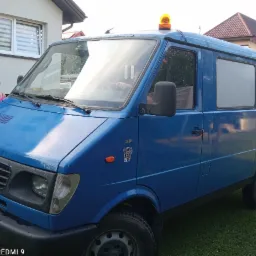 Niebieski samochód dostawczy z pomarańczowym światłem ostrzegawczym na dachu, zaparkowany na trawie przed domem jednorodzinnym.