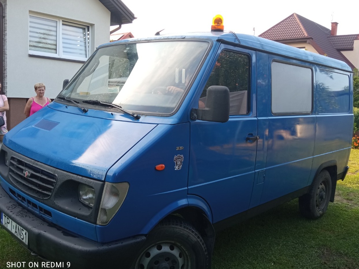 Niebieski samochód dostawczy z pomarańczowym światłem ostrzegawczym na dachu, zaparkowany na trawie przed domem jednorodzinnym.