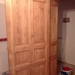 Odnowione, drewniane drzwi z czterema panelami, widoczne ślady po renowacji, obok stoi wiadro z farbą i pędzle, w tle grzejnik i fragment ściany z instalacją elektryczną.