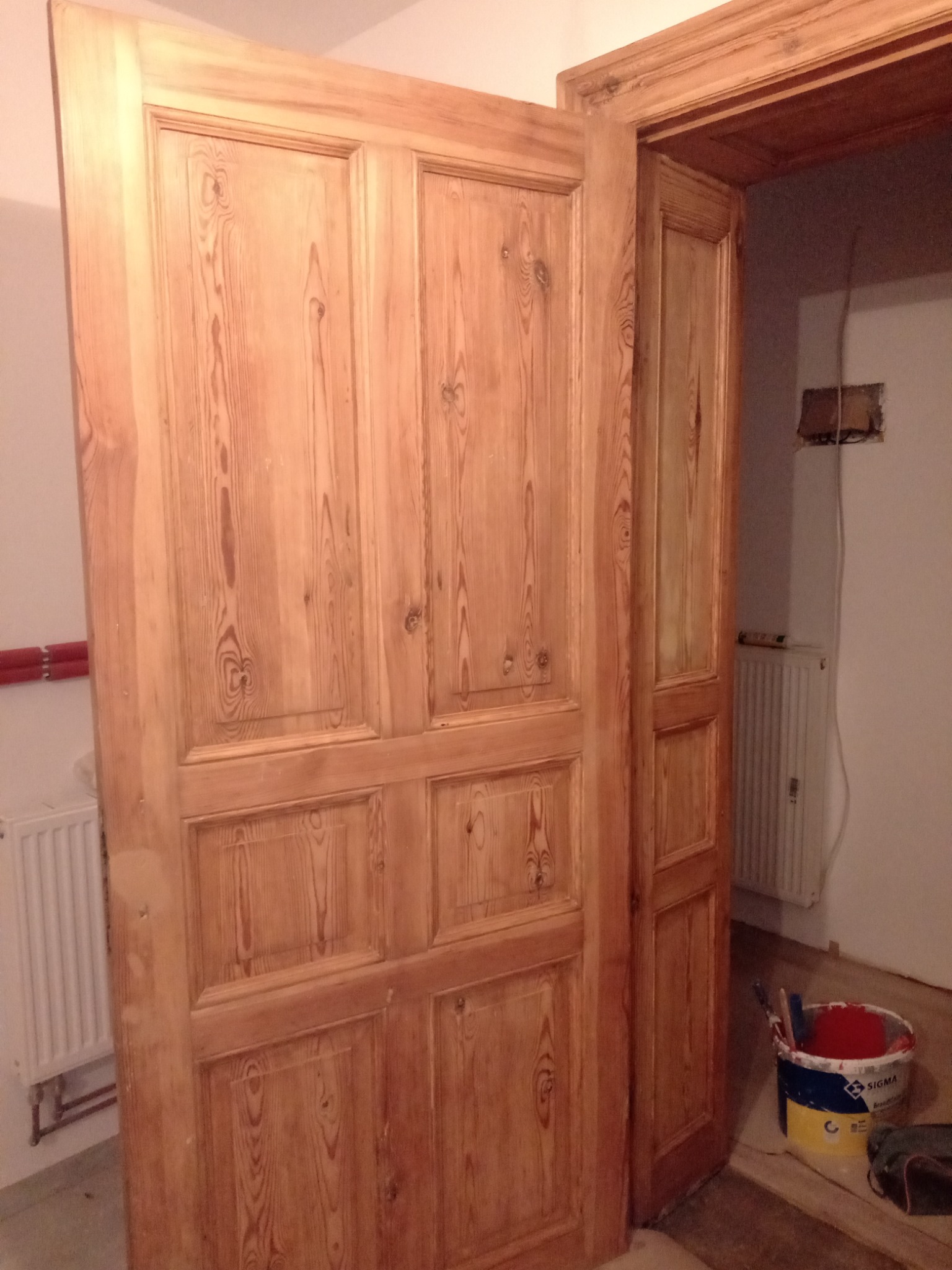 Odnowione, drewniane drzwi z czterema panelami, widoczne ślady po renowacji, obok stoi wiadro z farbą i pędzle, w tle grzejnik i fragment ściany z instalacją elektryczną.