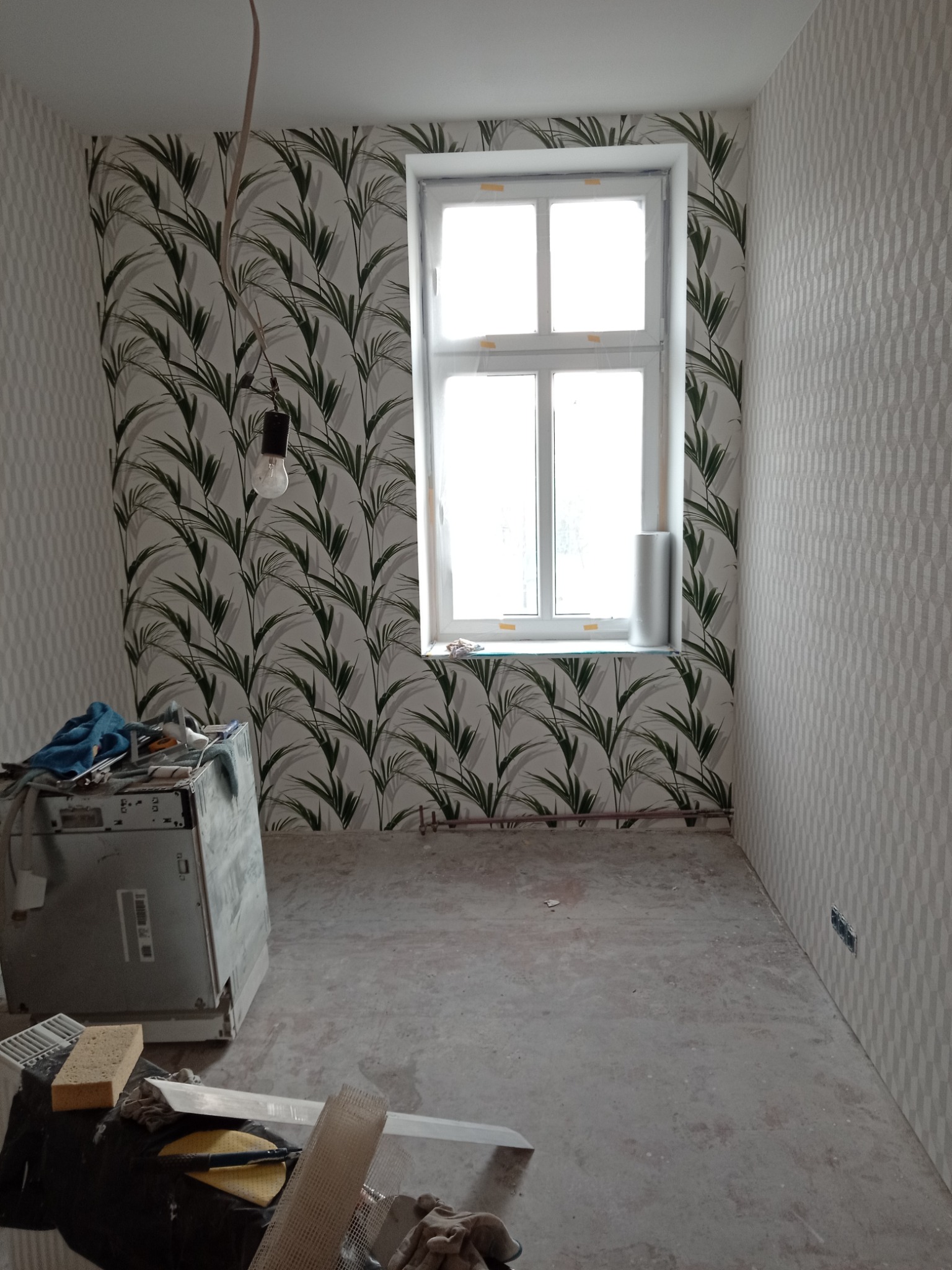 Pokój w trakcie prac wykończeniowych: ściany częściowo pokryte tapetą w motywy liści, druga ściana z geometrycznym wzorem, okno z folią ochronną, surowa podłoga, widoczne narzędzia i sprzęt malarski.