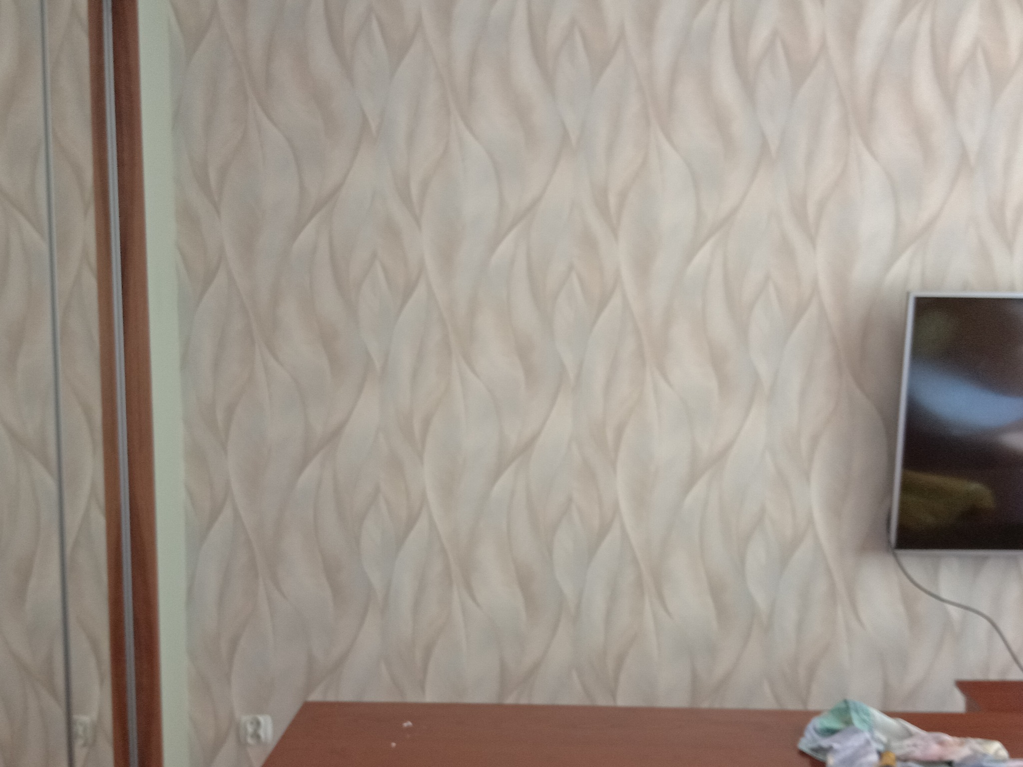 Ściana z tapetą o wzorze liści w odcieniach beżu, widoczny fragment szafy z lustrem po lewej i telewizor po prawej stronie, na stole leżą szmatki.
