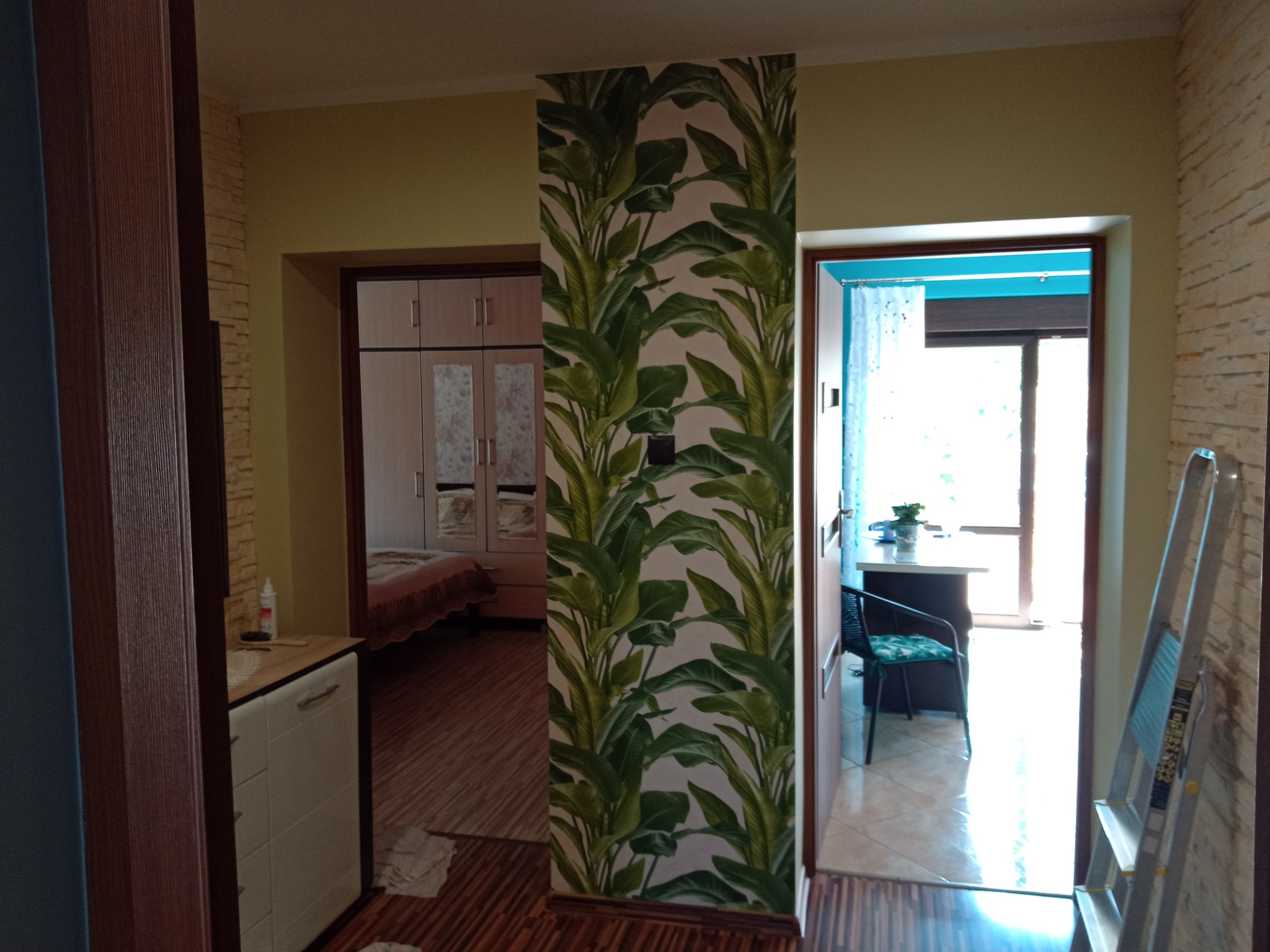 Fragment wnętrza po odświeżeniu: kolumna z tapetą w motywie tropikalnych liści pomiędzy pokojami, widoczna drabina malarska i fragmenty mebli w tle.