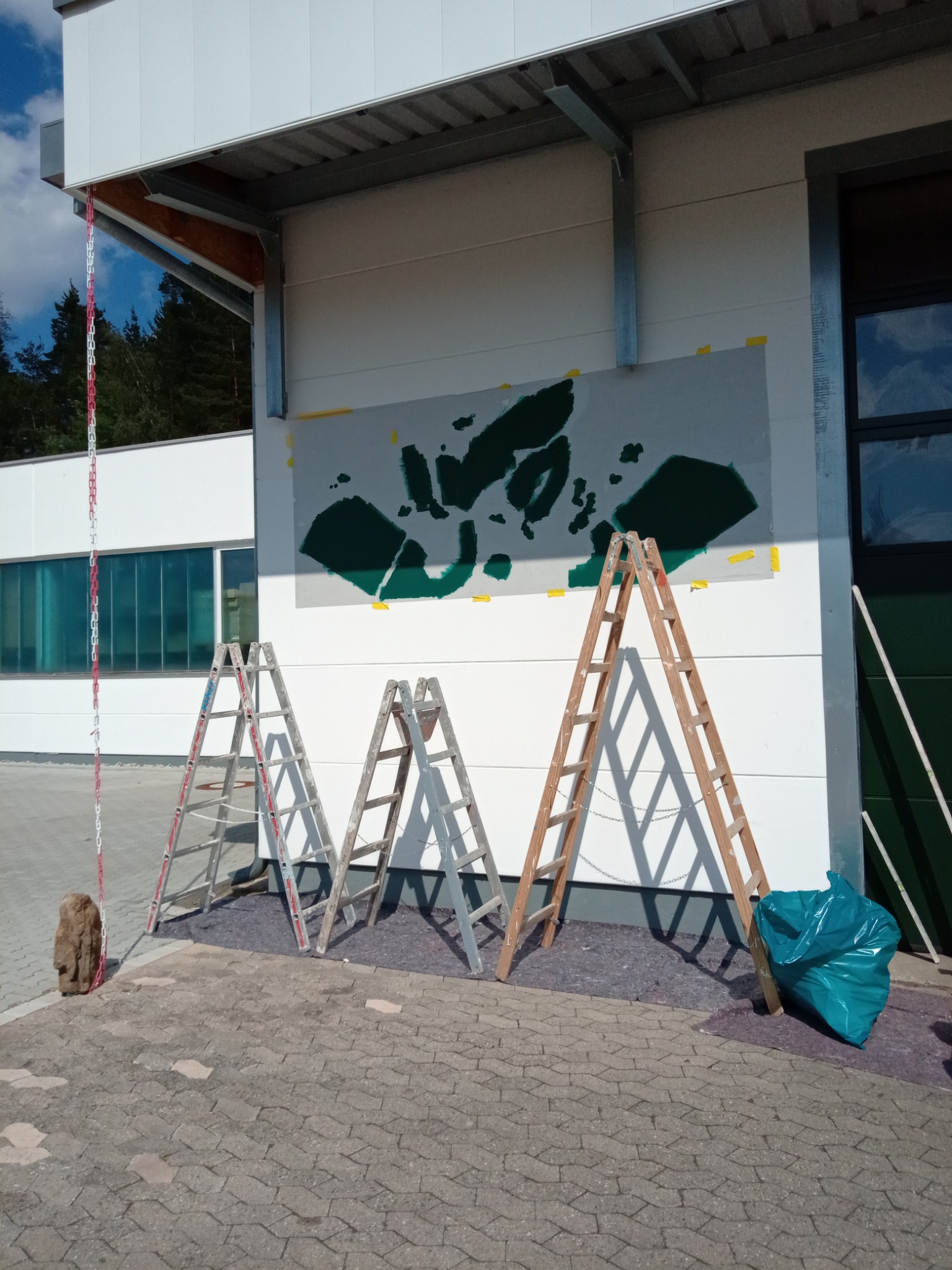 Malowanie wzoru na elewacji budynku przemysłowego, widoczne trzy drabiny, zabezpieczona folia i worek na odpady.