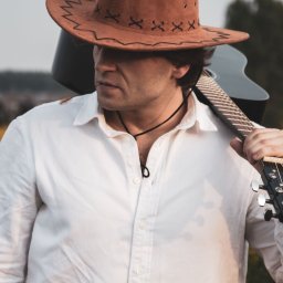 Marta Piątek Fotografia - Mężczyzna w kowbojskim kapeluszu i białej koszuli trzyma na ramieniu gitarę akustyczną, patrząc w lewo.