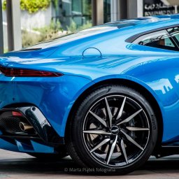 Marta Piątek Fotografia - Błękitny sportowy samochód Aston Martin, widok z tyłu, z wyraźnie widocznym kołem z aluminiowymi felgami i tablicą rejestracyjną z napisem TIMELESS. W tle nowoczesna architektura.