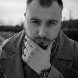 Marta Piątek Fotografia - Portret mężczyzny z brodą w płaszczu, zamyślony, dotyka dłonią brody, widoczny zegarek na nadgarstku, czarno-białe zdjęcie.