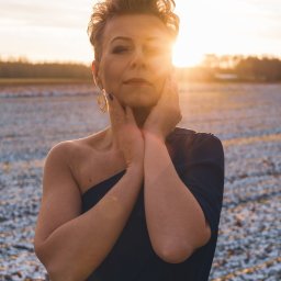 Marta Piątek Fotografia - Kobieta w granatowej sukni pozuje na zasypanym śniegiem polu, z zachodzącym słońcem za nią, oświetlającym jej twarz i włosy.