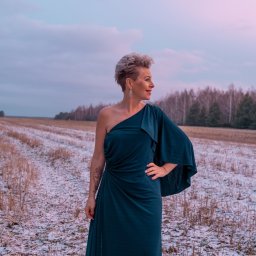 Marta Piątek Fotografia - Kobieta w szmaragdowej sukni na jedno ramię, z tatuażem na ramieniu, pozuje na zaśnieżonym polu w promieniach zachodzącego słońca, z lasem w tle.