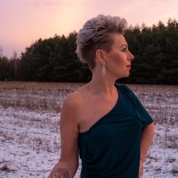 Marta Piątek Fotografia - Kobieta w ciemnozielonej sukni na tle zimowego krajobrazu o zmierzchu, widoczny tatuaż lwa i kwiatów na ramieniu, srebrne kolczyki i krótka fryzura.
