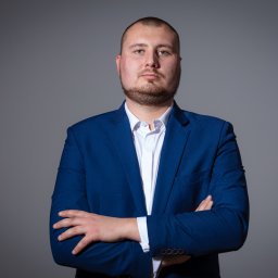 Marta Piątek Fotografia - Portret mężczyzny w niebieskim garniturze i białej koszuli, z założonymi rękami na szarym tle, wykonany przez fotografa Martę Piątek.