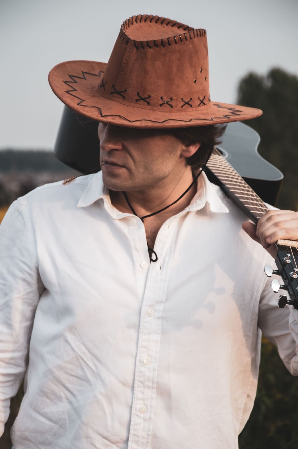 Mężczyzna w kowbojskim kapeluszu i białej koszuli trzyma na ramieniu gitarę akustyczną, patrząc w lewo.