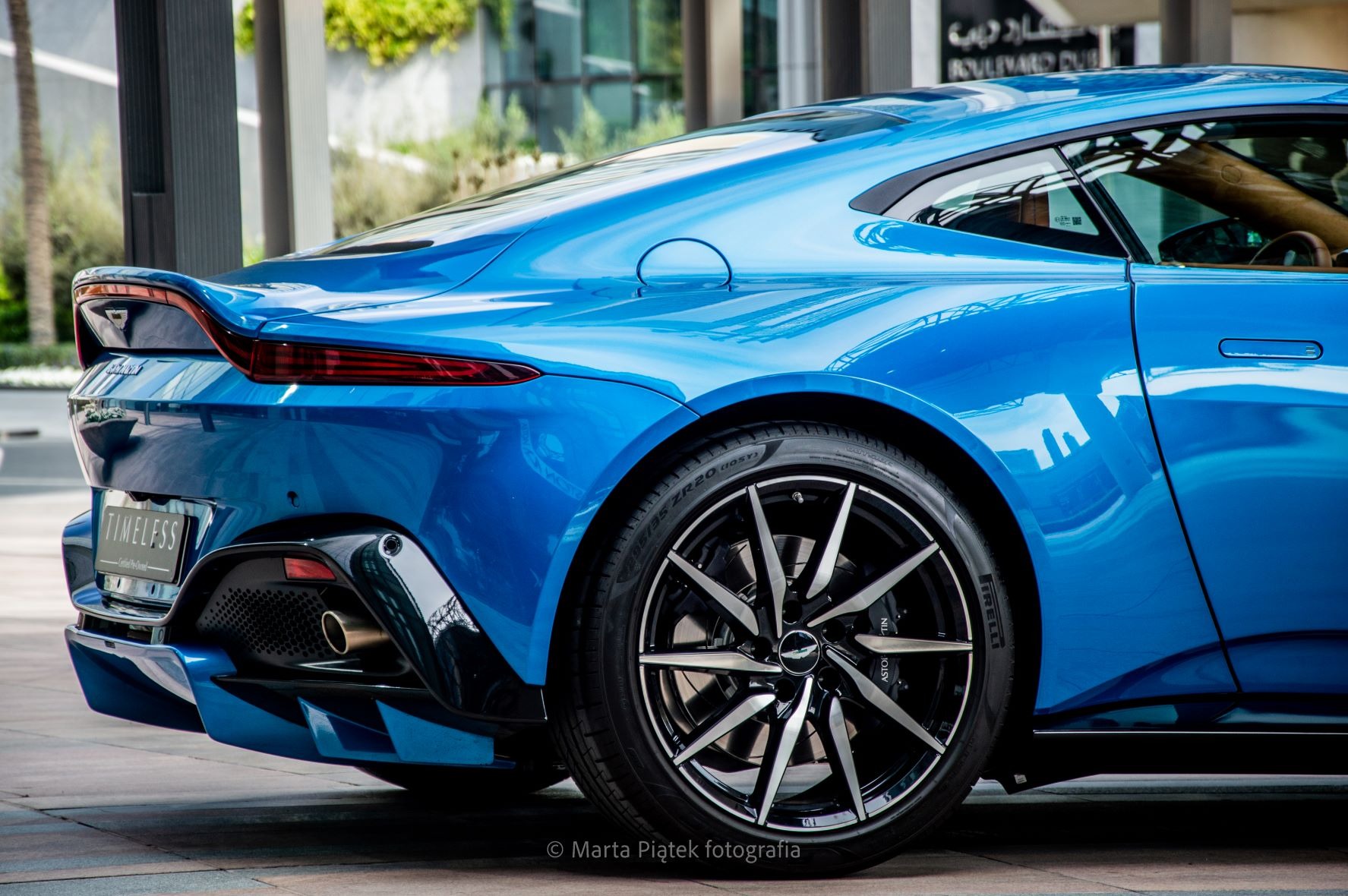 Błękitny sportowy samochód Aston Martin, widok z tyłu, z wyraźnie widocznym kołem z aluminiowymi felgami i tablicą rejestracyjną z napisem TIMELESS. W tle nowoczesna architektura.