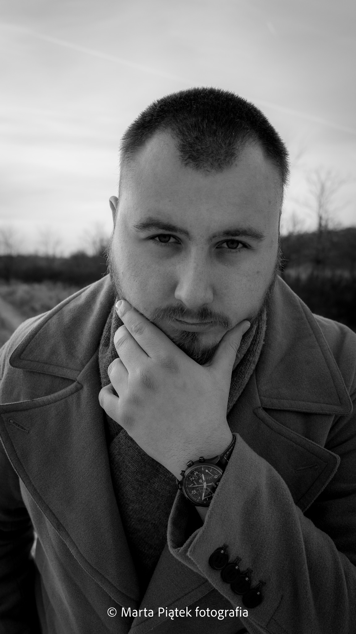 Portret mężczyzny z brodą w płaszczu, zamyślony, dotyka dłonią brody, widoczny zegarek na nadgarstku, czarno-białe zdjęcie.