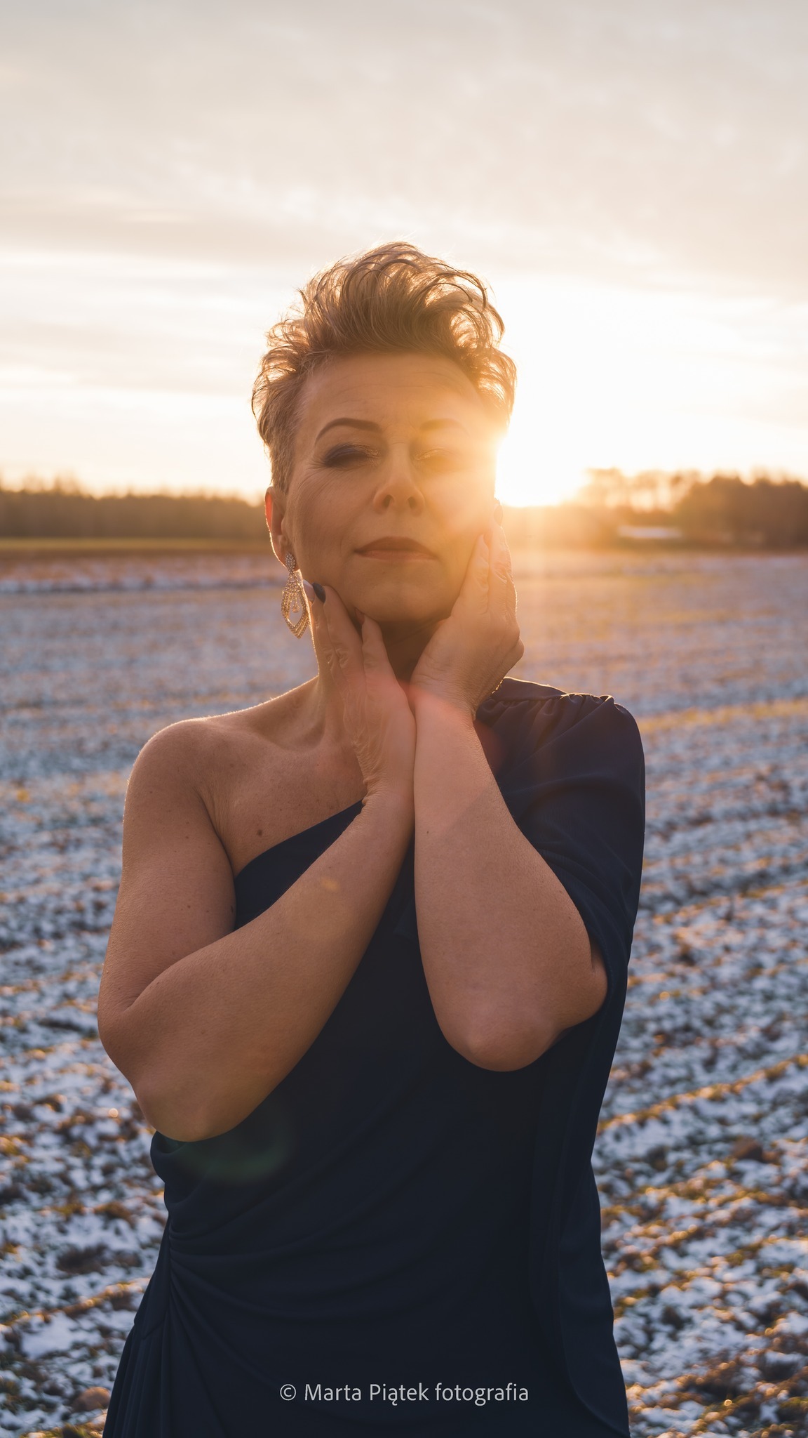 Kobieta w granatowej sukni pozuje na zasypanym śniegiem polu, z zachodzącym słońcem za nią, oświetlającym jej twarz i włosy.