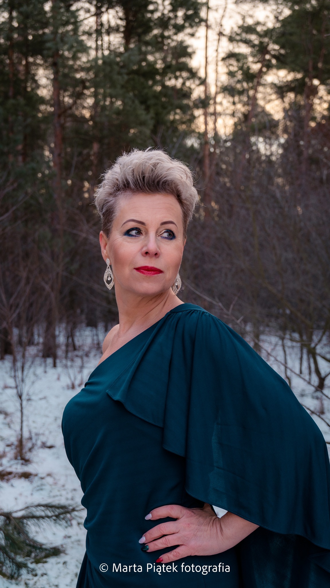 Kobieta w szmaragdowej sukni na tle zimowego lasu, pozująca z ręką na biodrze i spoglądająca w bok, z widoczną biżuterią.