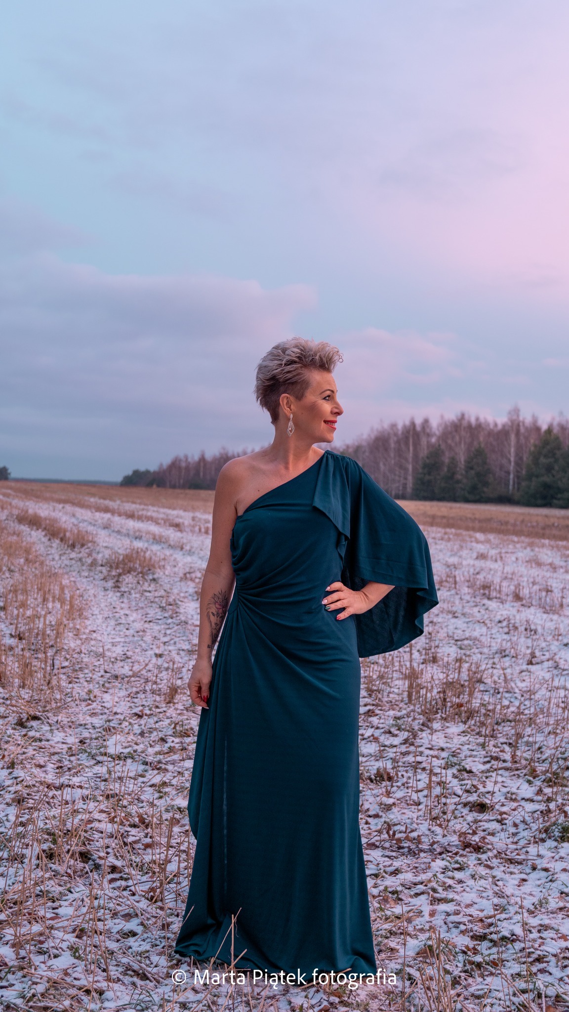 Kobieta w szmaragdowej sukni na jedno ramię, z tatuażem na ramieniu, pozuje na zaśnieżonym polu w promieniach zachodzącego słońca, z lasem w tle.