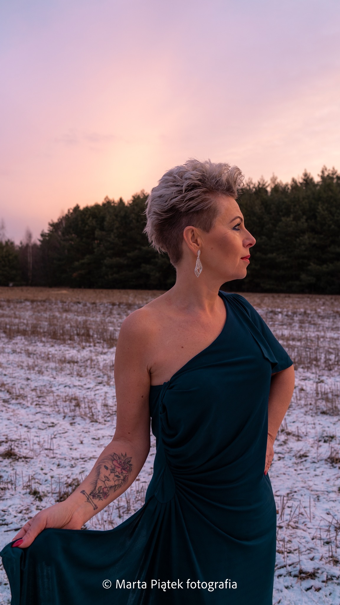 Kobieta w ciemnozielonej sukni na tle zimowego krajobrazu o zmierzchu, widoczny tatuaż lwa i kwiatów na ramieniu, srebrne kolczyki i krótka fryzura.