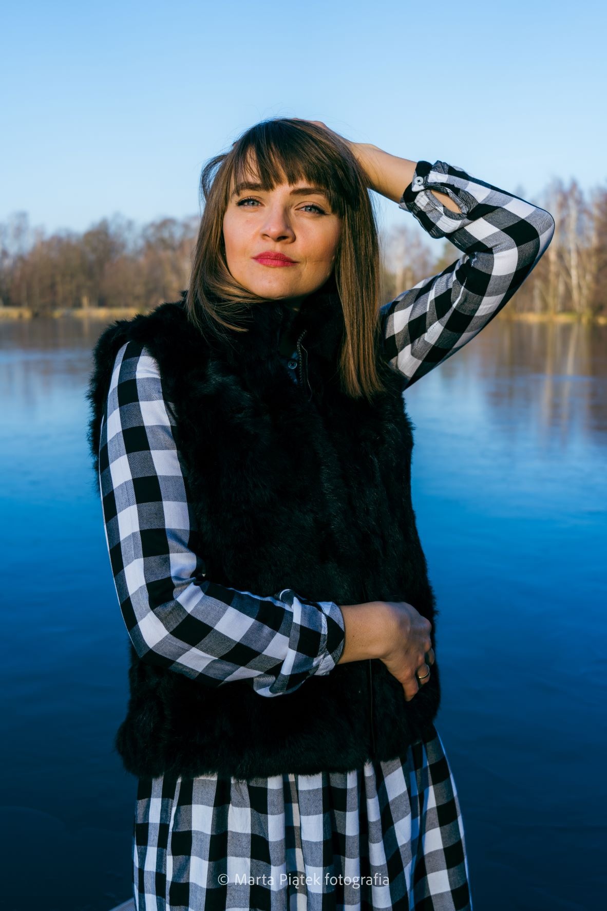 Portret kobiety w czarno-białej kraciastej sukience i futrzanej kamizelce na tle jeziora i drzew, z ręką uniesioną nad głową.