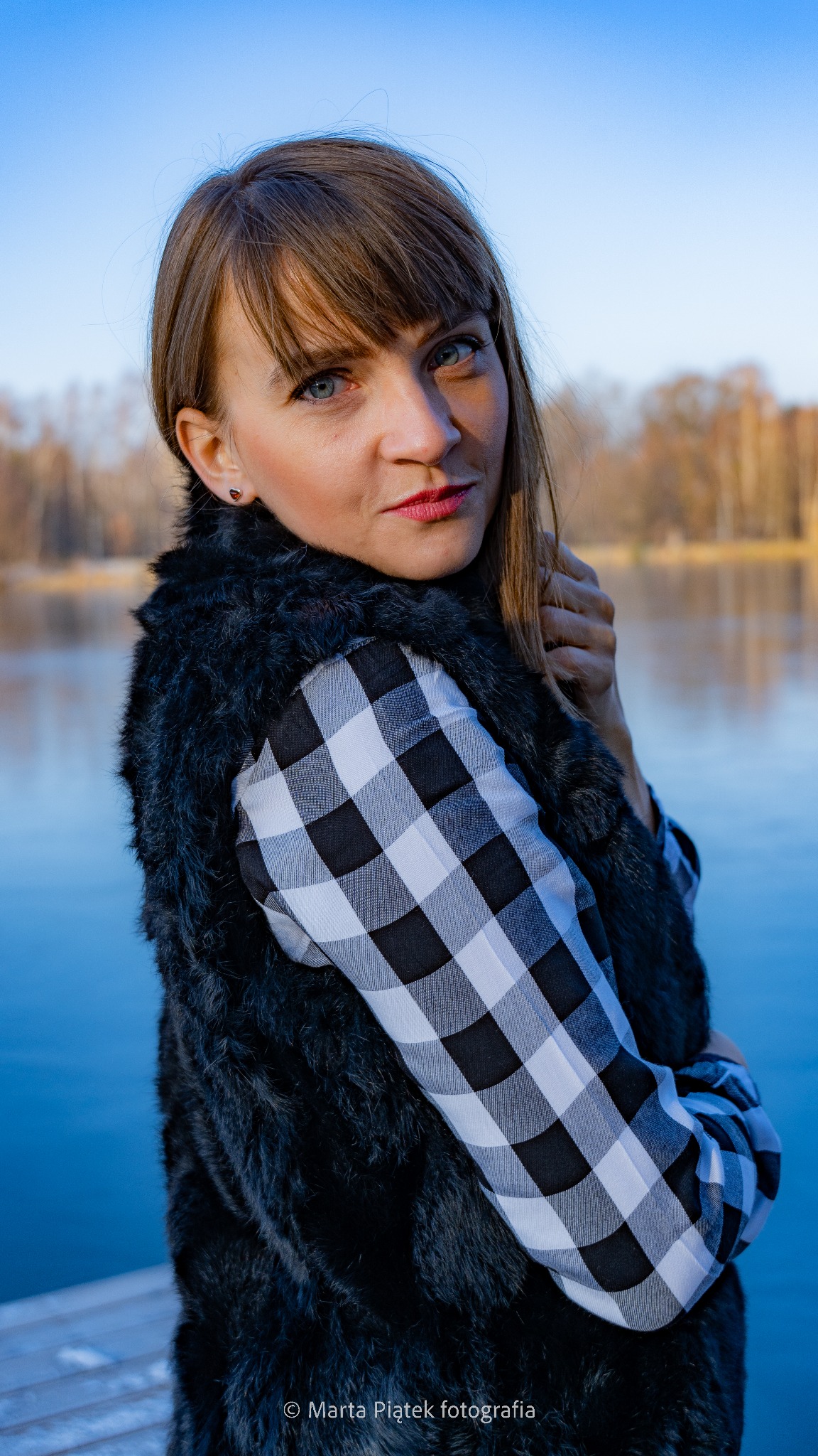 Portret kobiety w futrzanej kamizelce i koszuli w kratę na tle jeziora, z delikatnym uśmiechem i spojrzeniem w obiektyw.