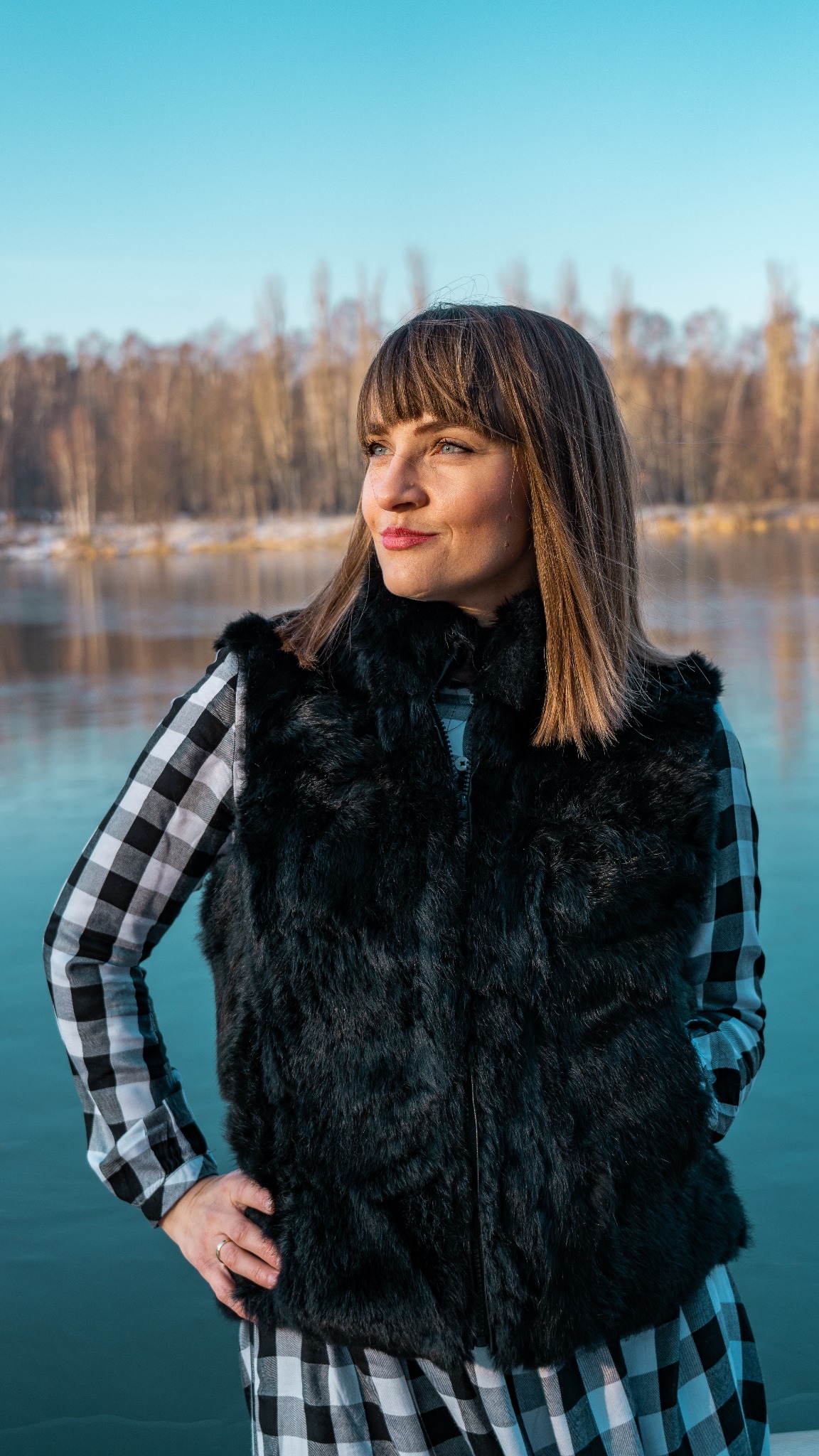 Kobieta w czarnej futrzanej kamizelce i kraciastej koszuli pozuje na tle jeziora i lasu w zimowym krajobrazie, spoglądając w lewo.