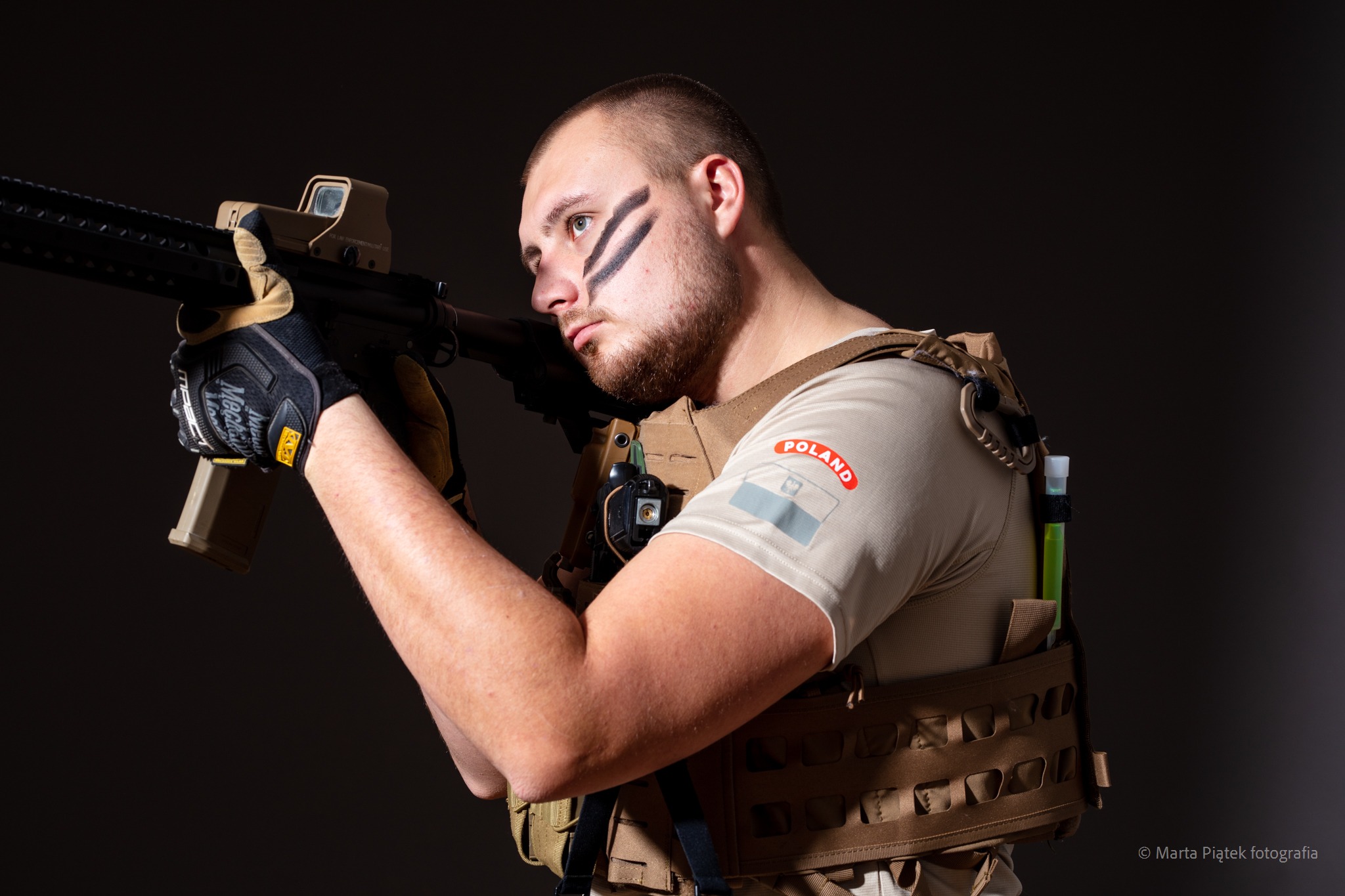 Mężczyzna w kamizelce taktycznej, z flagą Polski na ramieniu i czarnymi paskami na twarzy, celuje z karabinu w ciemnym otoczeniu.