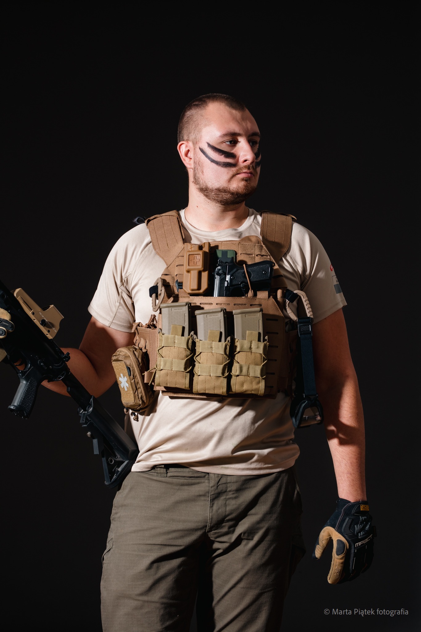 Portret mężczyzny w kamizelce taktycznej, z farbami wojennymi na twarzy, trzymającego karabin na tle ciemnego studia.