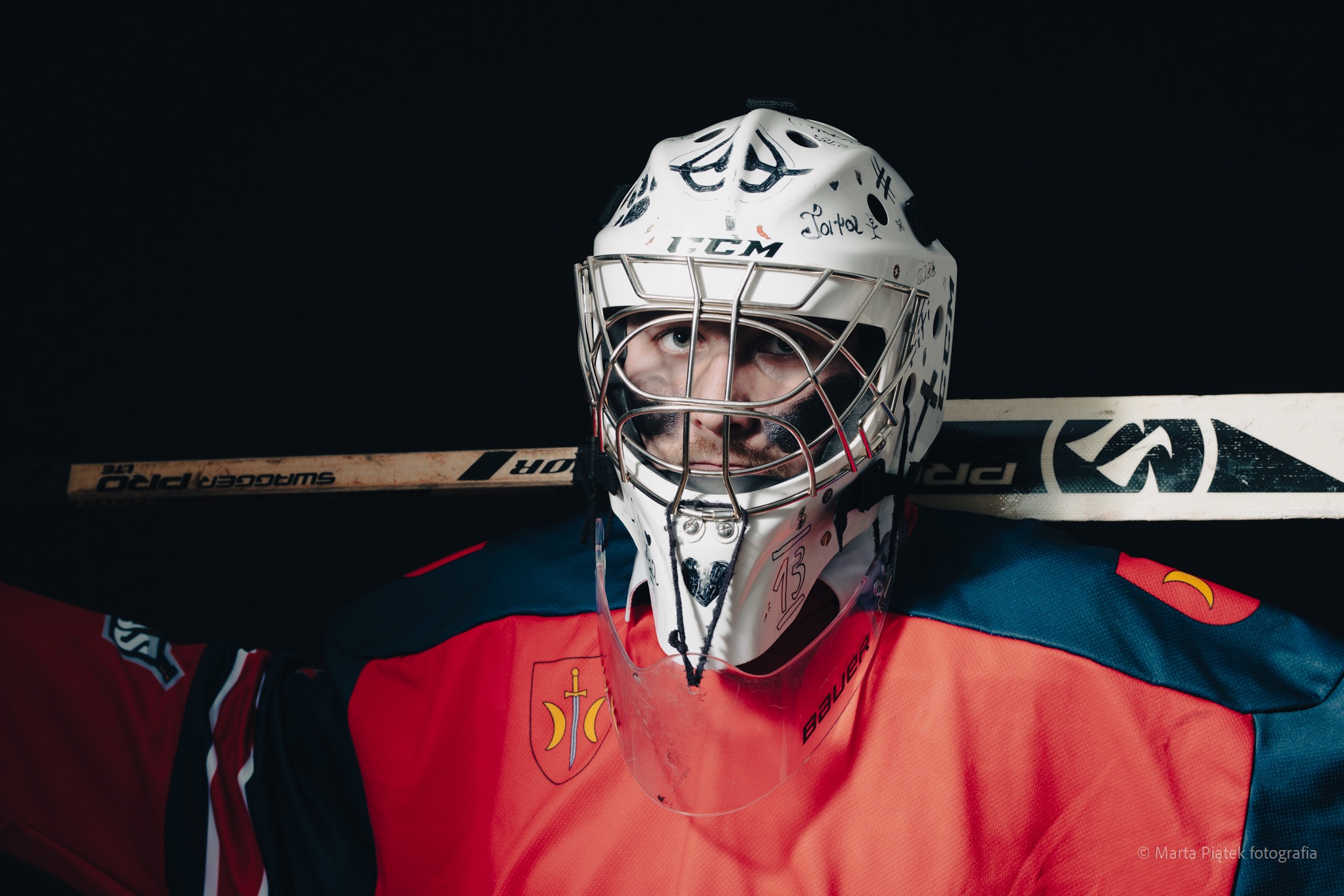 Portret hokejowego bramkarza w masce z niestandardowymi rysunkami, w koszulce z herbem, z kijami hokejowymi w tle.